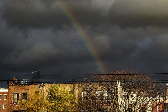 A rainbow over the rooftops of some buildings visible through dark clouds.